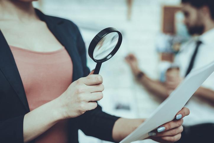 Detectives working in Private Detective Agency. Woman is Looking on Paper with Magnifying Glass. Man is Standing at Clues Map on Background.