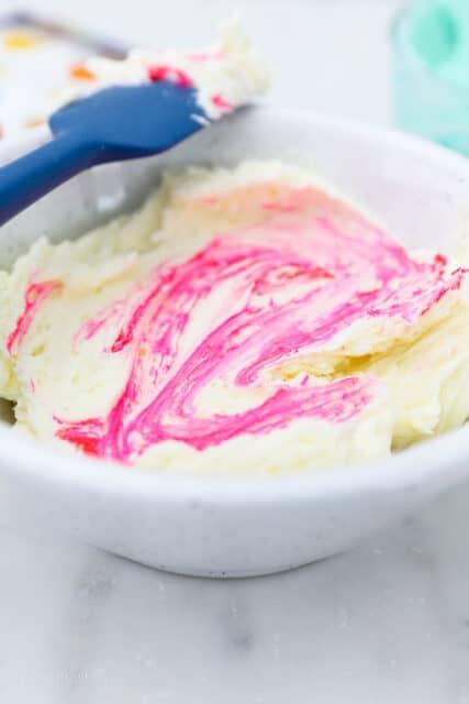 Overhead view of vanilla buttercream frosting in a bowl, next to two bottles of gel food dye and a color chart.