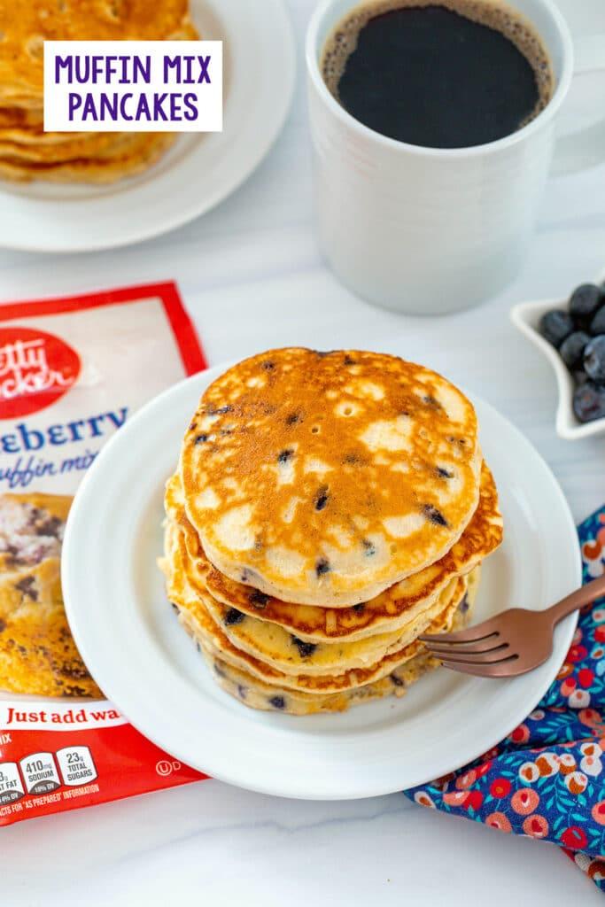 Overhead view of a stack of blueberry muffin mix pancakes with muffin mix bag, second plate of pancakes, and cup of coffee in background with recipe title at top