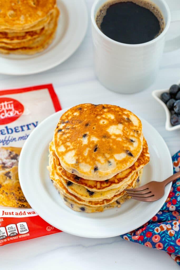 Overhead view of a stack of blueberry muffin mix pancakes with muffin mix bag, second plate of pancakes, and cup of coffee in background