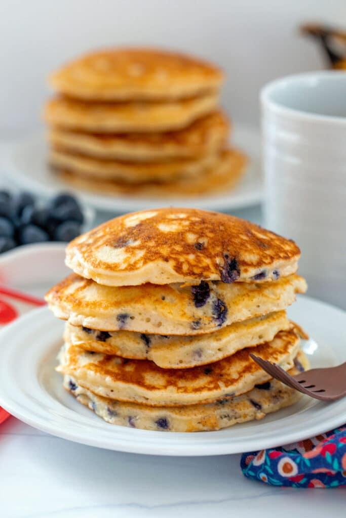 Stack of blueberry muffin mix pancakes on plate with fork and more pancakes in background