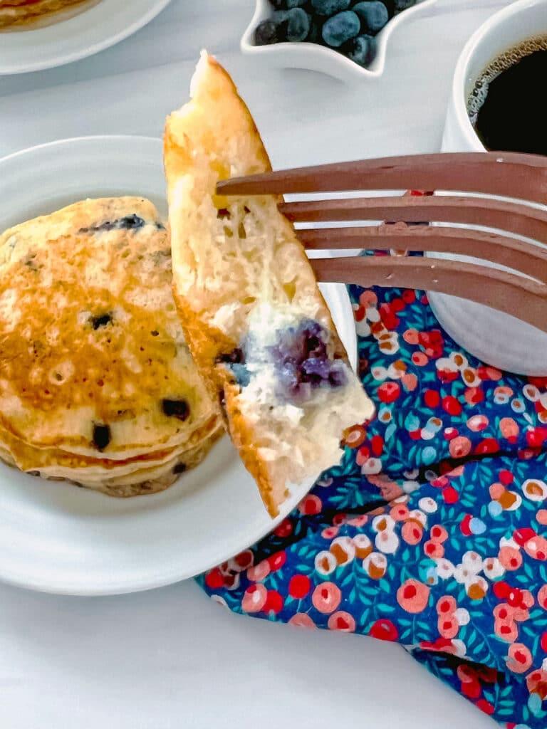 Bite of blueberry muffin mix pancake on a fork with pancakes in background