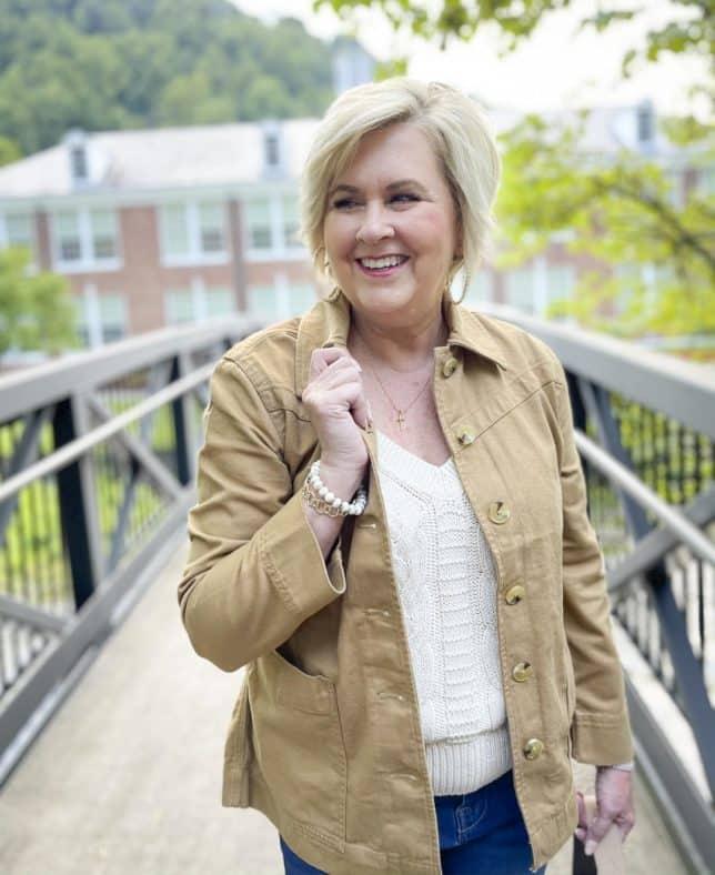 Over 40 Fashion Blogger, Tania Stephens, is standing on a bridge in flare jeans, a neutral handbag, and a camel colored jacket