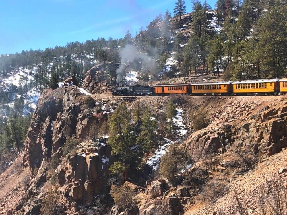 a yellow/orange train along the side of a cliff drop off, smoke coming out the front is a great activity from Durango to Pagosa Springs