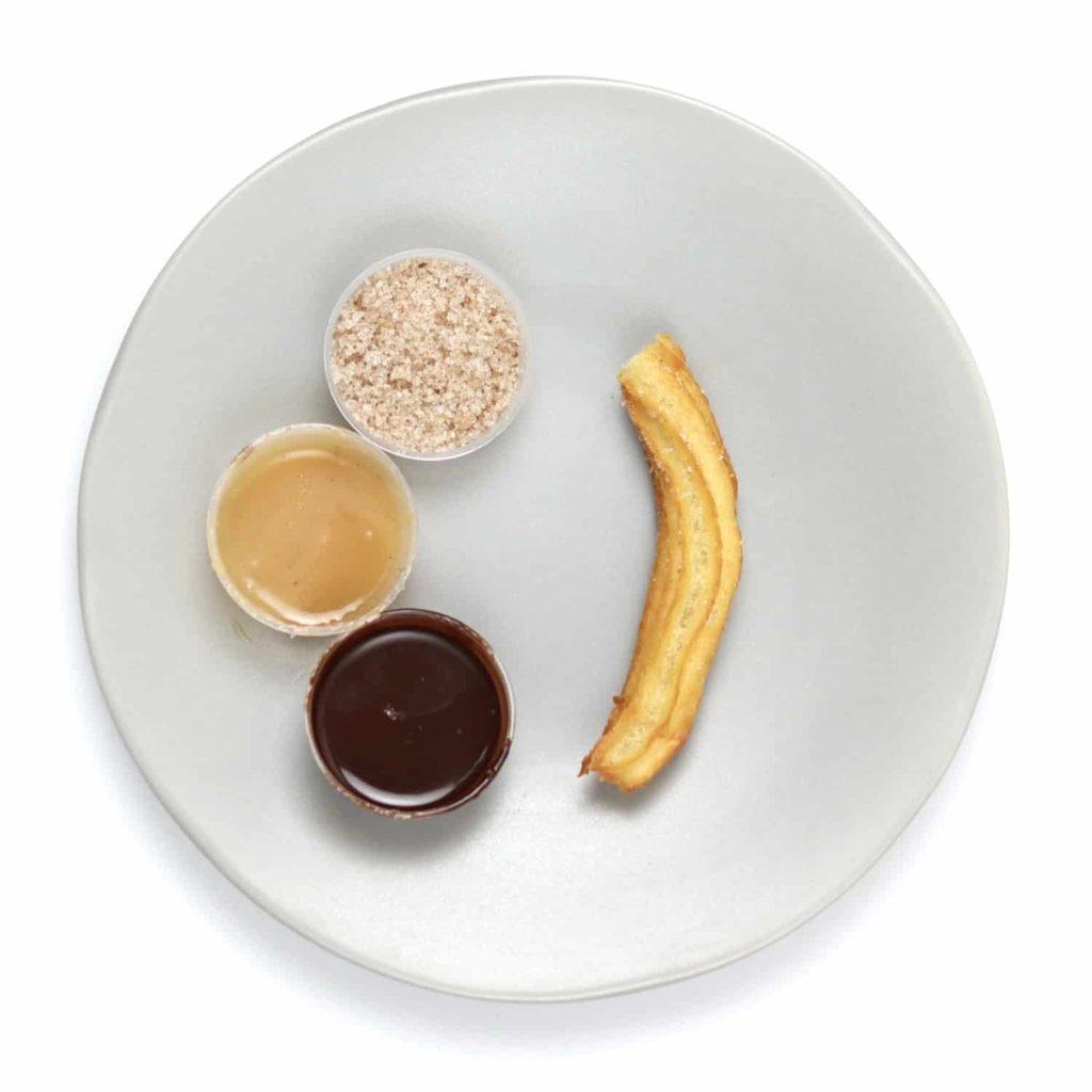 Single churro on a small plate alongside chocolate sauce and cinnamon sugar