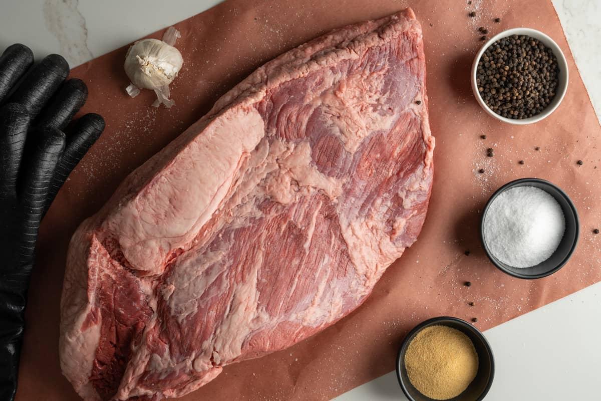 untrimmed brisket surrounded by garlic powder, salt, pepper corns, and rubber gloves