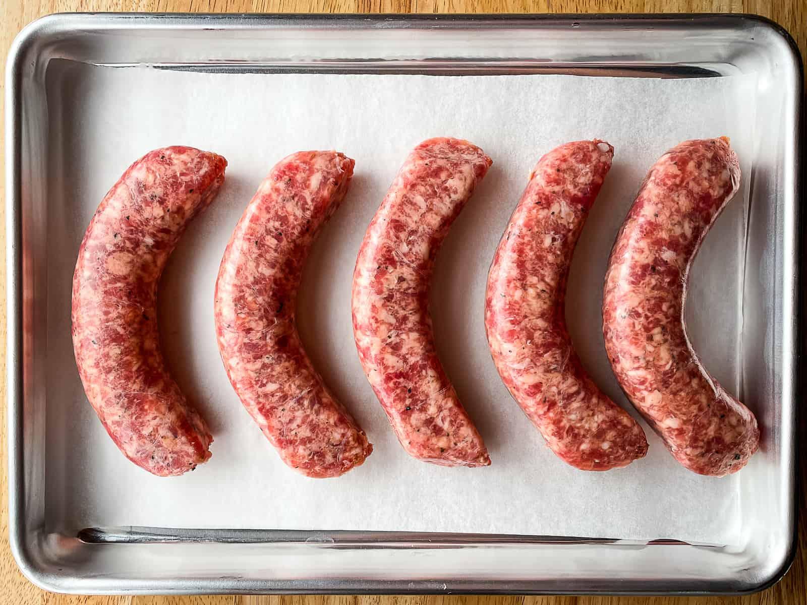 Five raw Italian sausage on a parchment lined baking sheet.