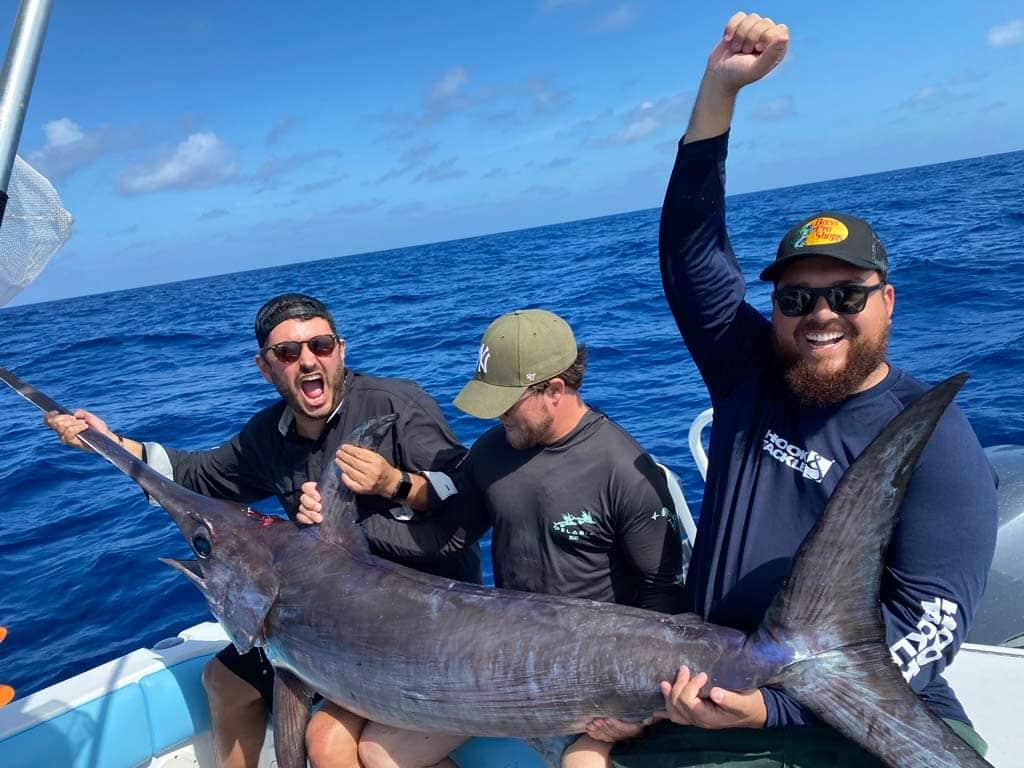 A group of three anglers sitting on the back deck of a boat during with a huge Swordfish that they caught daytime fishing lying across their laps.