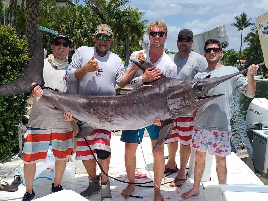 Five anglers standing on a docked boat, all of them holding a big Swordfish they caught daytime fishing.