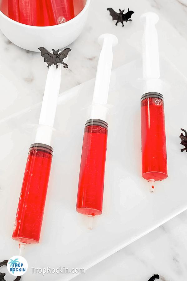 Red Jello shots in Syringes on a serving tray with little plastic black bats for decor.