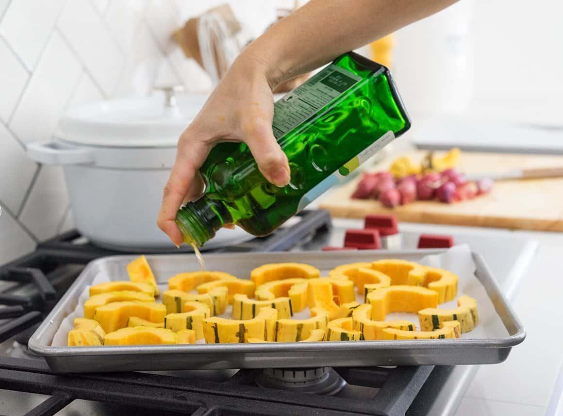 Hand drizzling olive oil over chopped vegetables on a baking sheet