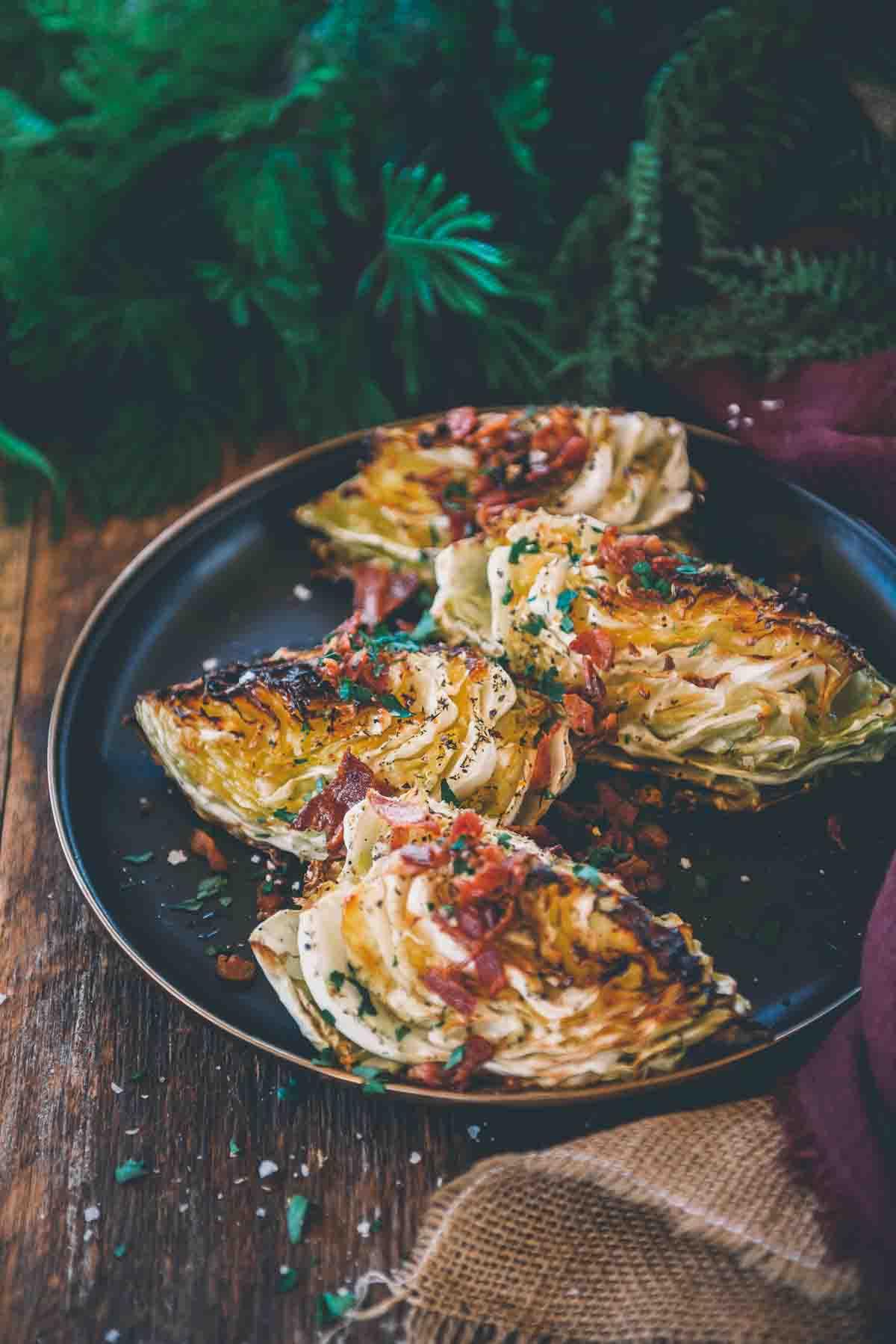 Black platter with 4 wedges of smoked cabbage with crispy golden brown edges garnished with crumbled bacon.