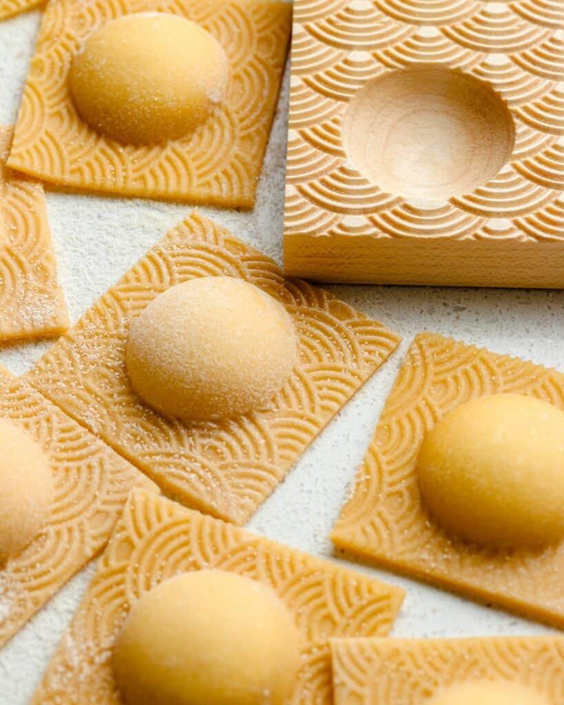 Ravioli on a bench top with the ravioli mold next to them.