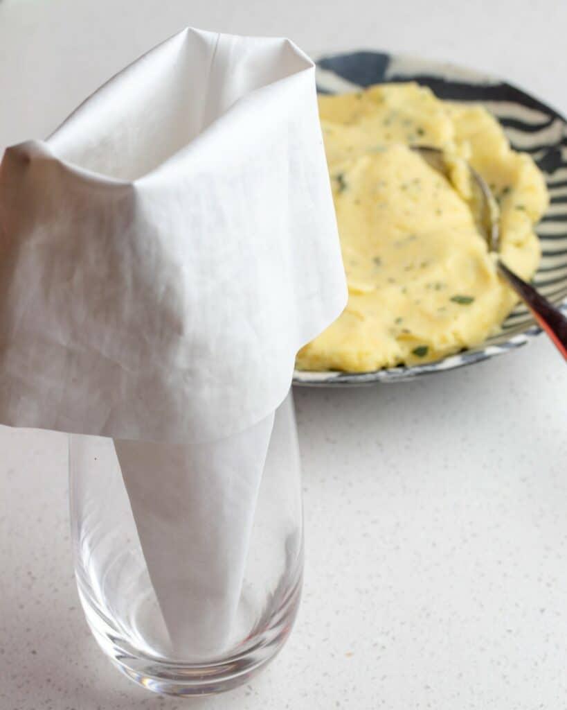 A glass with a piping bag upright in it and a bowl of ravioli filling in background.