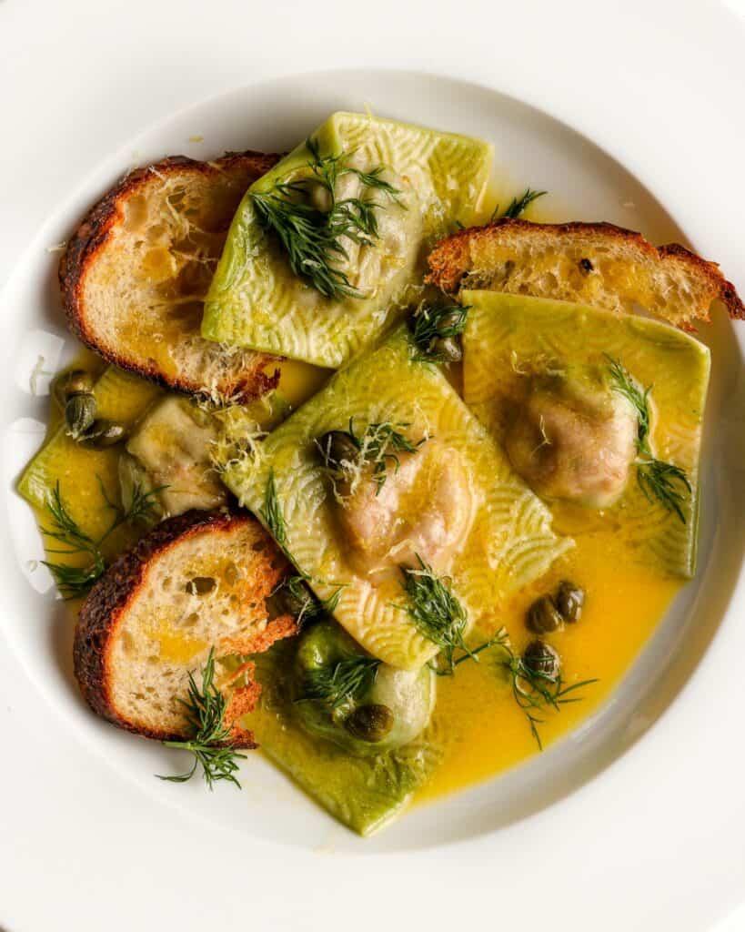 White plate with cooked ravioli in a yellow butter sauce with sourdough croutons, capers and dill.